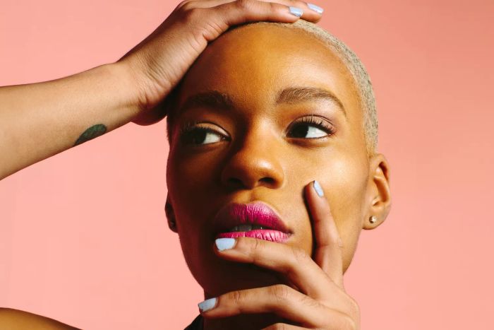 A stunning young woman with her hands gently resting on her face, sporting blue nails, gazing sideways against a pink studio backdrop