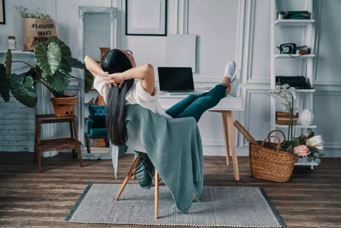 A young woman seated at her home office with her hands behind her head, looking relaxed.