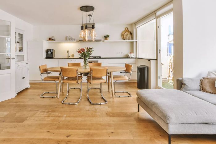 A modern minimalist-style studio apartment with a light design, featuring a wooden table and chairs in the dining area, seamlessly connecting to an open kitchen and living room with white walls and parquet flooring.