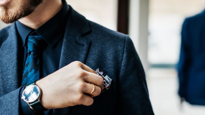 A close-up image of a man carefully placing a handkerchief into his breast pocket.