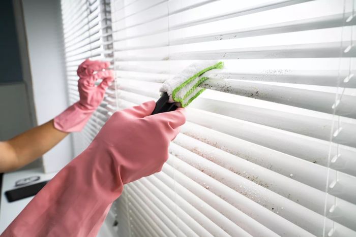 A person using a duster to clean white window blinds from accumulated dirt