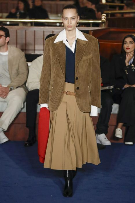 A model showcases a layered outfit, with a collar peeking out from beneath a tan blazer on the runway.