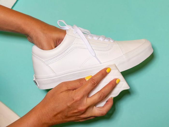 A person cleaning a white canvas shoe with a magic eraser, set against a teal background.