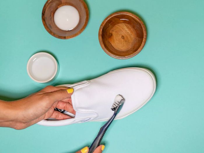 A person using a toothbrush to clean a white shoe, placed on a teal surface.