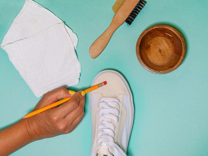 A person is cleaning a white suede shoe using a pencil eraser, surrounded by a white cloth, brush, and bowl, all displayed against a teal background.