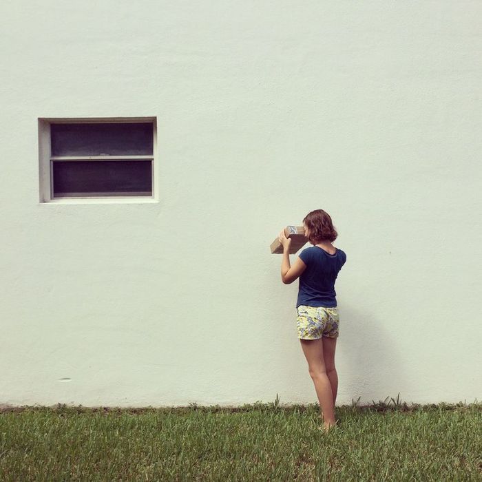 A young girl gazes at a blank white wall while peering into a handmade cardboard pinhole camera.