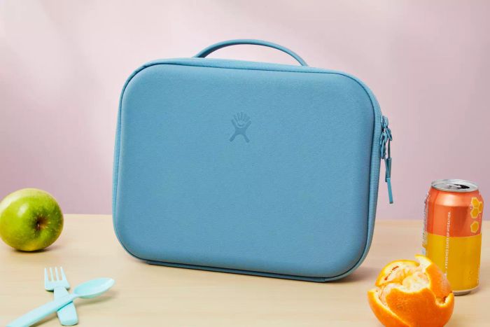 Hydro Flask Large Insulated Lunch Box resting on a desk next to fruit, cutlery, and a can of drink