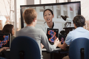 Students with tablets watching a female teacher on a monitor.