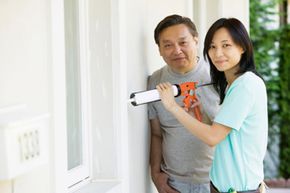 An Asian man and woman are sealing cracks on the house exterior with a caulk gun.