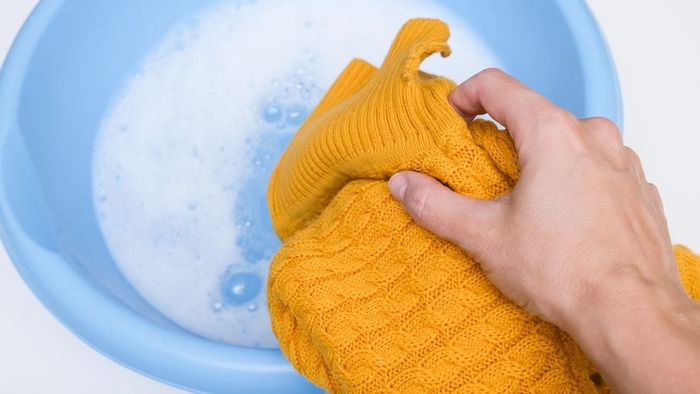 A hand gently placing a soft mustard yellow cashmere sweater into a basin filled with sudsy water.