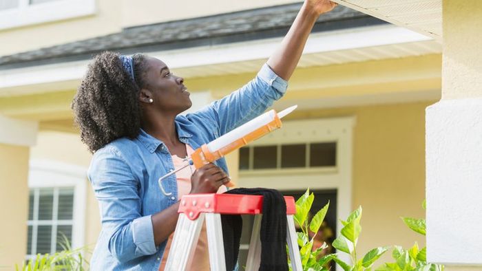 A young woman stands on a ladder outside her home, using a caulking gun to fix a leak.