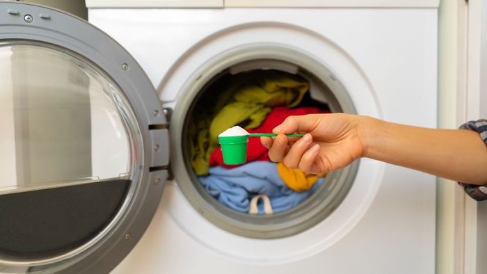 A woman holds a cap filled with washing powder in front of a washing machine, where clothes are tumbling inside.