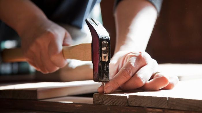 A hand using a hammer to drive a nail into a wooden surface.
