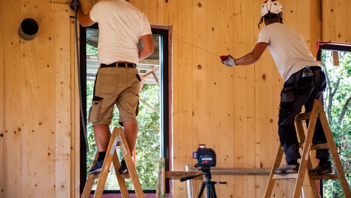 Two construction workers handling a ladder at a building site within a wooden house structure.