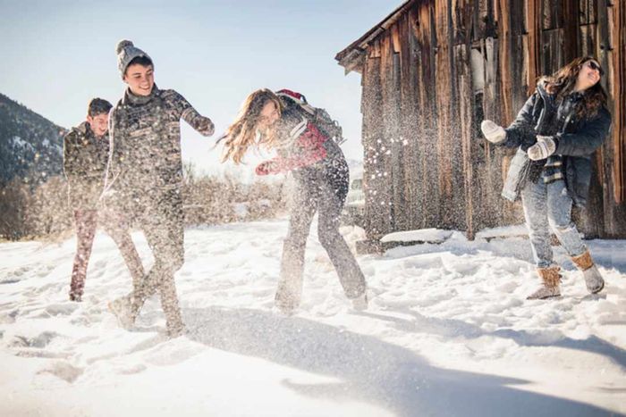 Four individuals dressed in winter attire engage in a snowball fight next to a rustic barn.