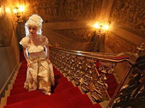 A woman in vintage attire and a wig ascending a golden staircase with red velvet lining.