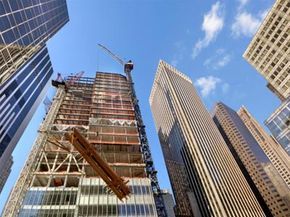 A skyscraper rises with steel beams being lifted by a crane, viewed from a low angle