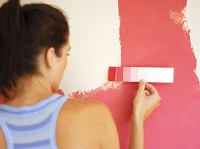 A woman is comparing various pink paint chips to the pink wall to find the closest match in color.