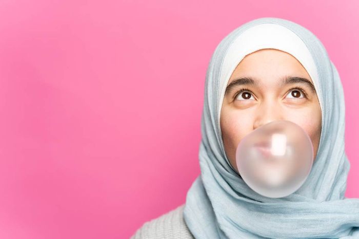A young woman blowing a bubble while chewing gum, set against a pink backdrop.