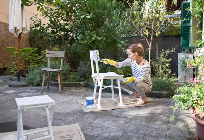 A woman sitting on a patio, applying white paint to a chair