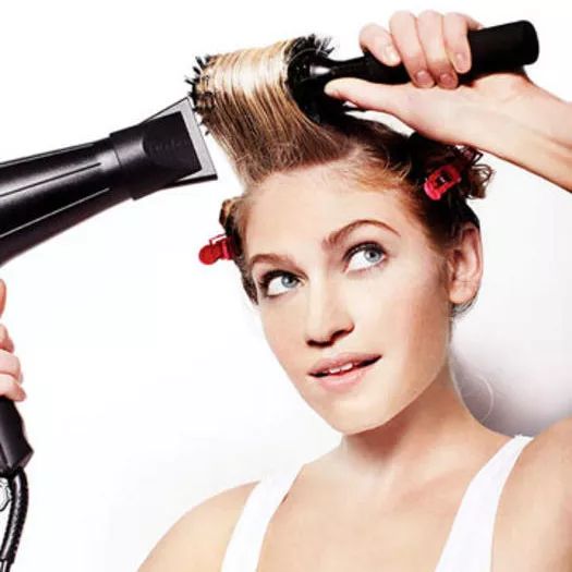 A person blow-drying their hair in sections, using a round brush for the crown area.