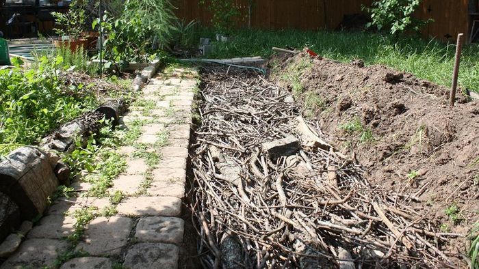 A hugelkultur garden bed made of organic matter and branches neatly arranged beside an ancient stone pathway.
