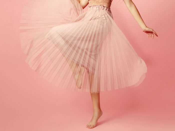Image of a woman from the waist down, wearing a pleated skirt, with one knee raised as she balances on one foot.