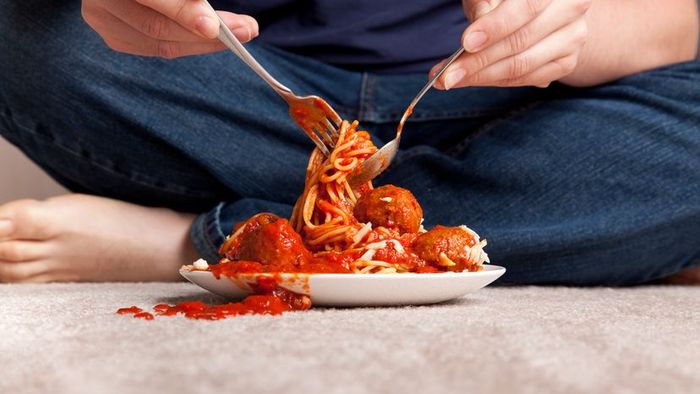 A person enjoying spaghetti while sauce drips onto the carpet.