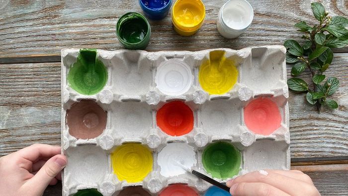 Children's hands create colorful paintings using an egg carton as a base on a wooden surface.