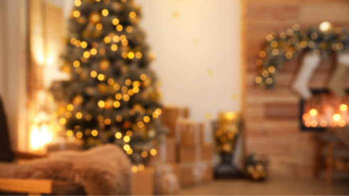 A festive Christmas tree adorned with golden ornaments and lights, surrounded by gifts and complementary holiday decorations, captured in a softly blurred image.