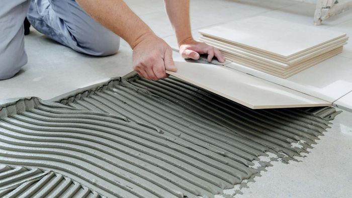 A worker kneels down to lay large white floor tiles onto pre-applied adhesive