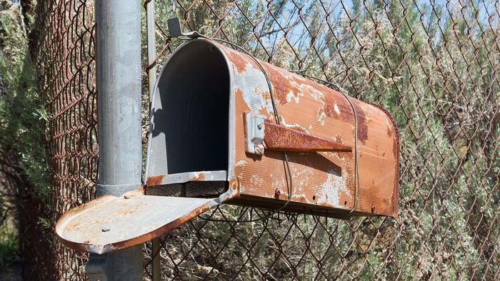 A corroded mailbox dangling from a fence