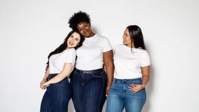 A group of women dressed in white t-shirts and blue jeans pose against a pristine white backdrop.
