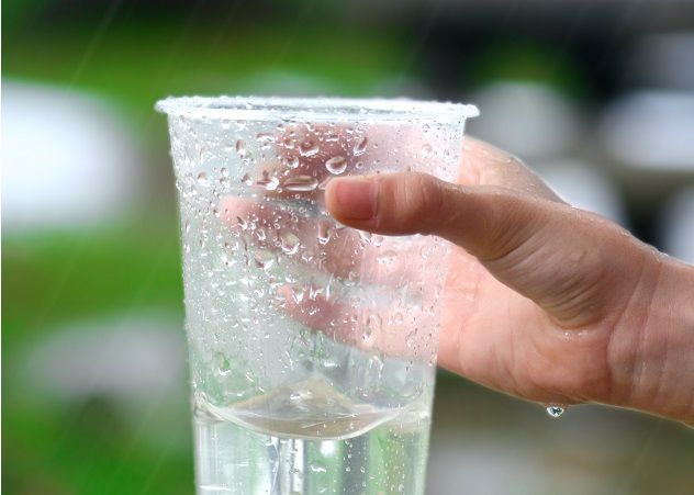 A child's hand grips a plastic cup, collecting rainwater as it falls.