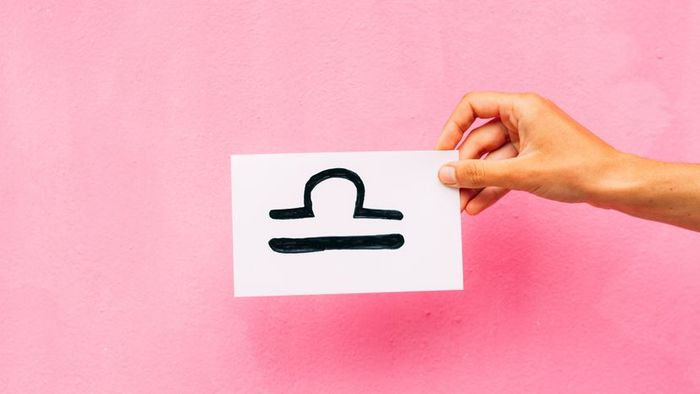 A hand holding a sheet of paper featuring the Libra zodiac symbol, set against a soft pink backdrop.