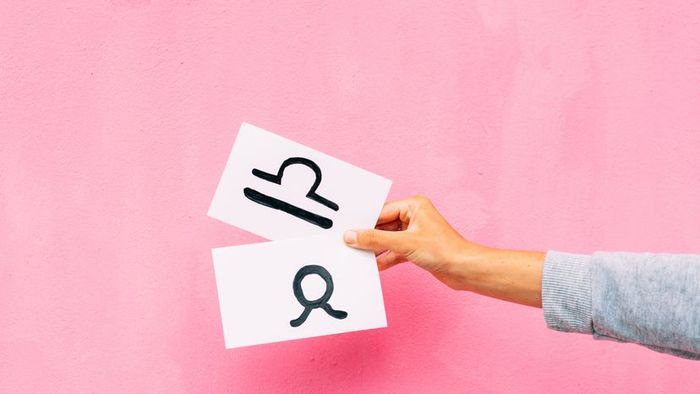 A hand clutching a white sheet adorned with the Libra symbol, set against a soft pink backdrop.