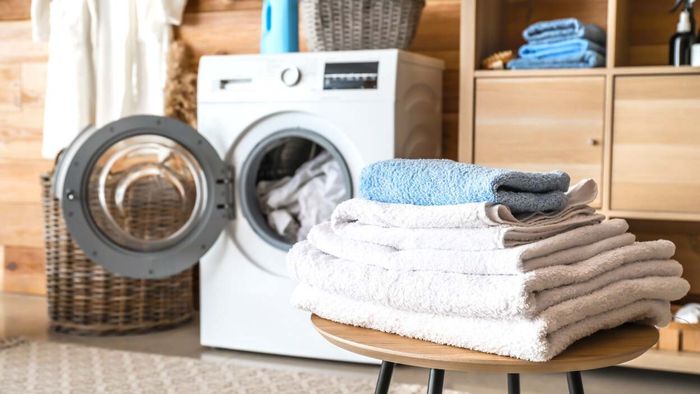 A stack of neatly folded towels rests on a stool positioned in front of an open front-loading washing machine.