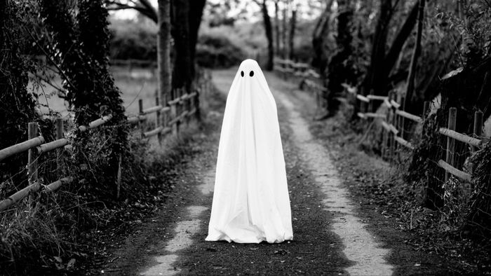A ghost draped in a white sheet stands on a quiet countryside trail.