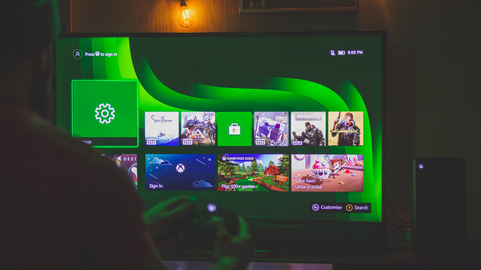 An image depicting a person seated with their back to the viewer, engaging in an Xbox game on a large TV screen in a room with subdued lighting