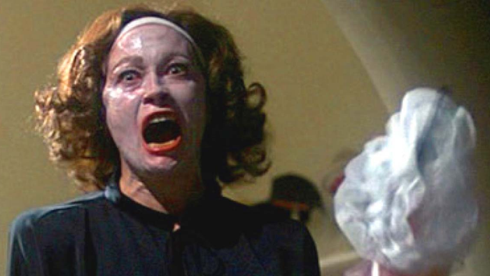 An image of Faye Dunaway, her face smeared with cold cream while wielding a wire hanger, from the movie Mommie Dearest