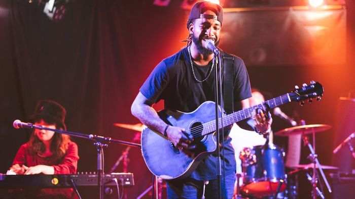 A Black male guitarist performing on an acoustic guitar during a live performance.
