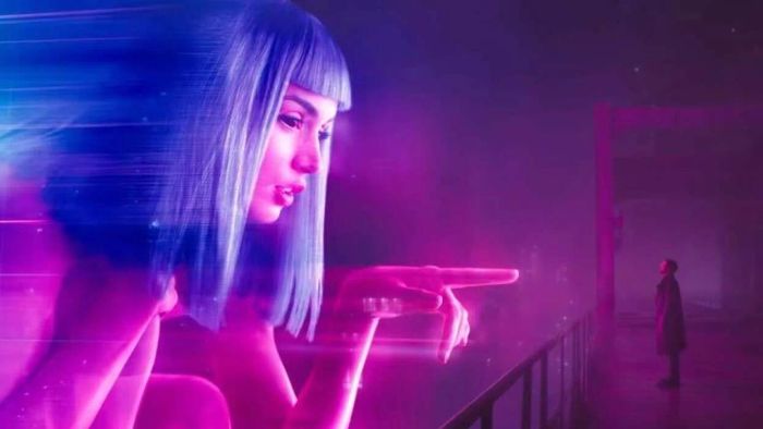 A still from Blade Runner 2049 showing a massive blue-haired holographic woman gesturing toward Ryan Gosling's character on a shadowy balcony