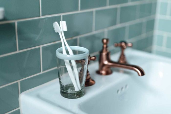 Two toothbrushes placed in a glass beside a copper faucet on a sink.