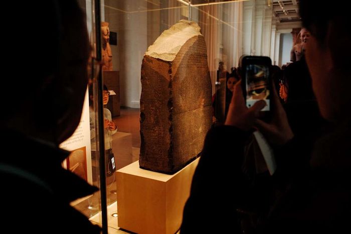 The Rosetta Stone is now showcased in a glass enclosure at the British Museum in London, depicted here with shadowed visitors in the background.