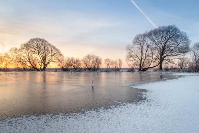 A serene winter pond, blanketed with a thin sheet of ice and snow, surrounded by trees as the sun sets in the background.