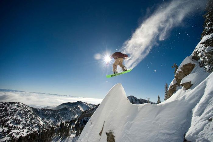 A snowboarder leaps off a cliff, leaving a trail of powdery snow in the air, set against a bright blue sky and sun.