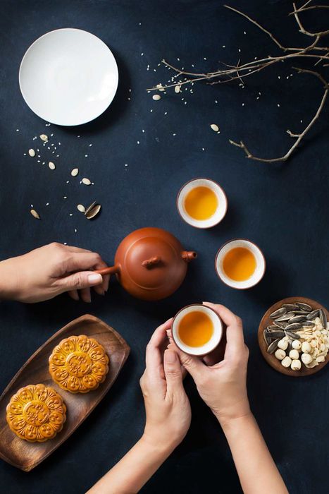 A flat-lay scene of a Mid-Autumn Festival tea gathering, featuring food, drinks, and a hand pouring tea.