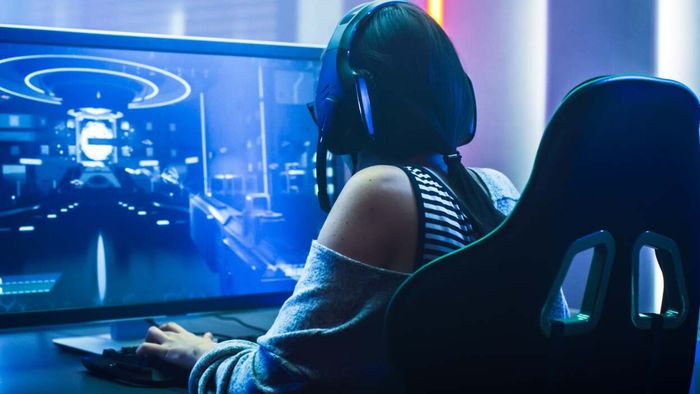 A woman is seated at a computer, engaged in a game while wearing a headset