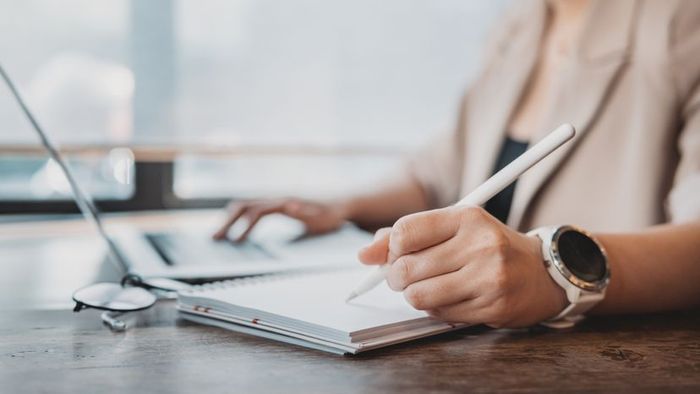 A woman jotting down notes on a notepad while working on her laptop.