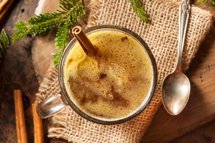 A top-down view of a glass mug filled with hot spiced rum, placed on a burlap cloth alongside a spoon, pine sprigs, and cinnamon sticks.